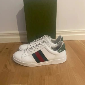 Gucci ace - Säljer ett par klassiska vita Gucci sneakers med gröna detaljer på hälen och de ikoniska röd-gröna ränderna på sidan. Skorna har snörning och platt sula, perfekt för en stilren och trendig look.