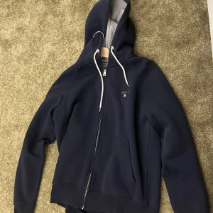 Mörkblå hoodie med dragkedja från Gant - Säljer en mörkblå hoodie från Gant i storlek L. Tröjan har dragkedja, huva med vita snören och två fickor framtill. Insidan av huvan är grå. Perfekt för en avslappnad stil. Hör gärna av er för fler frågor. 
