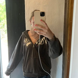 Mörkgrå hoodie med tryck från Divided - Säljer en mörkgrå hoodie från Divided med dragkedja och stort beige tryck framtill. Tröjan har huva, fickor och långa ärmar. Perfekt för en avslappnad stil och passar till jeans eller leggings. Den är i stl xxs men den är lite over saize