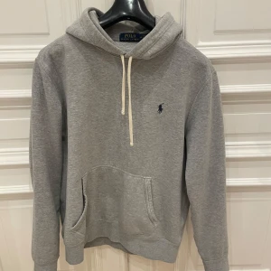 Grå hoodie från Polo Ralph Lauren storlek M - Säljer en stilren grå hoodie från Polo Ralph Lauren med klassisk logga på bröstet. Tröjan har en justerbar huva med vita snören och en praktisk magficka. Perfekt för en avslappnad look. Nästan ny skick kan gå ner i pris vid snabb affär bara att skriva vid funderingar.