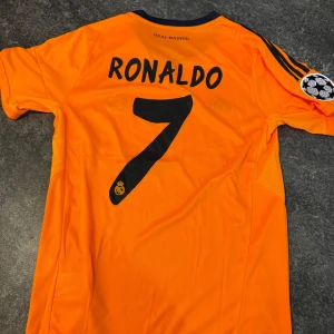 Real Madrid Ronaldo fotbollströja - Säljer en orange Real Madrid fotbollströja från Adidas med Ronaldo 7 på ryggen. Tröjan har svarta detaljer, klubbmärke och Champions League-patch på ärmen. Fly Emirates tryck på bröstet och Climacool-material.