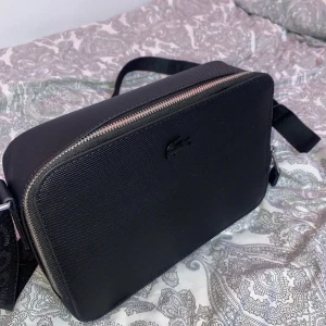 Svart axelväska från Lacoste - Snygg svart axelväska från Lacoste med dragkedja upptill. Väskan har en justerbar axelrem och en stilren design med Lacoste-loggan på framsidan. Perfekt för att bära dina nödvändigheter med stil.