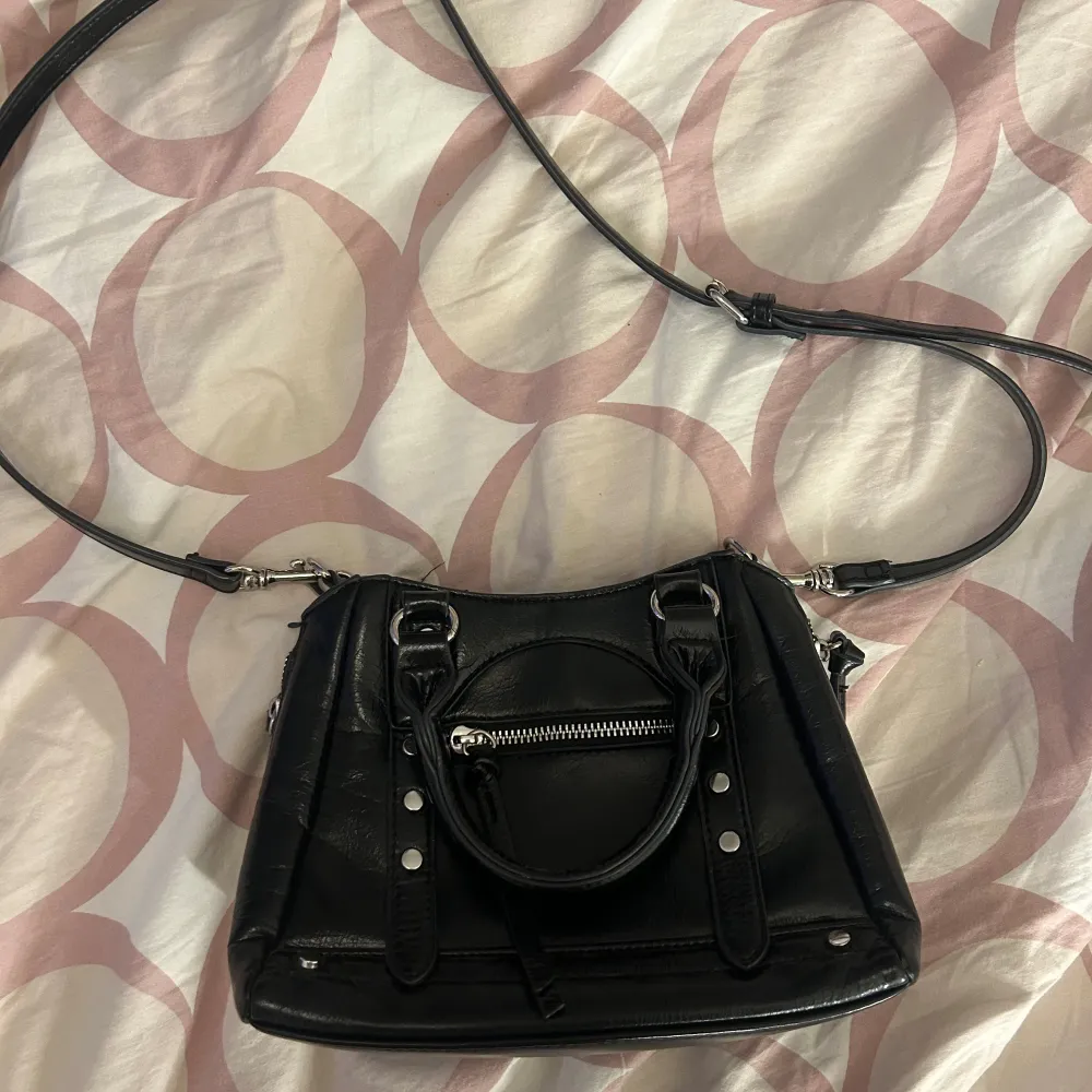 Säljer en svart axelväska med både handtag och justerbar axelrem. Väskan har silverfärgade detaljer, dragkedja framtill och dekorativa nitar. Perfekt storlek för det viktigaste och passar till många olika outfits.. Laukut & Käsilaukut.