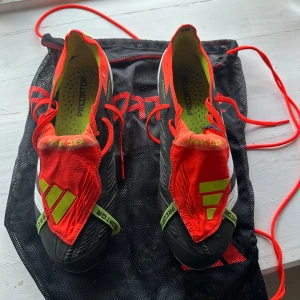Adidas Predator fotbollsskor i svart och röd - Säljer ett par Adidas Predator fotbollsskor i svart med röda och neongula detaljer. Skorna har snörning och en modern design med mönstrad ovandel och genomskinlig sula med dobbar. Perfekta för dig som vill sticka ut på planen!