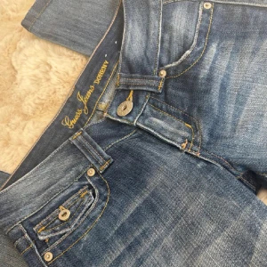 Blå jeans från Guess - Snygga blå jeans från Guess med klassisk femficksdesign och knappdetaljer.jag har köpt dom här på plick men dom var tyvärr försmå så jag behöver sälja dom igen. Dom har små slitningar längst ner vid föten men det syns inte alls när dom är på. Skickar gärna fler bilder och diskuterar priset🥰