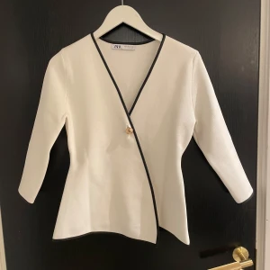 Vit omlottopp från Zara - Säljer en stilren vit omlottopp från Zara med svarta kanter och en dekorativ knappdetalj. Toppen har trekvartsärmar och en elegant passform. Aldrig använd