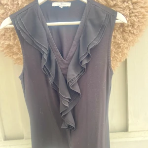 Svart ärmlös topp med volang från Flash Woman - Svart ärmlös topp från Flash Woman med  v-ringning och dekorativa volanger längs ringningen framtill. toppen har en enkel, rak passform och är tillverkad i ett mjukt tyg som känns bekvämt mot huden. SKRIV INNAN KÖP!❤️