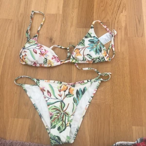 Blommig bikini med knyt -  Nypris var 350kr och den är oanvänd🩷 Säljer en snygg bikini med blommigt mönster i grönt, gult, rosa och orange på vit botten. Överdelen har smala axelband och knyt i ryggen, underdelen har smala sidor. Perfekt för sommaren!