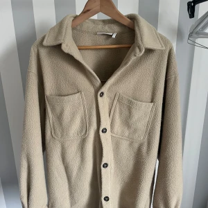 Beige fleeceovershirt från Weekday - Säljer en beige overshirt från Weekday i mjuk fleece med två stora bröstfickor och knappar framtill. Perfekt att slänga på sig över en t-shirt för en avslappnad look. Klassisk krage och lång ärm.