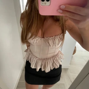 Beige korsettopp med volang - Säljer en beige korsettopp med volangdetaljer och markerad midja. Toppen har en romantisk känsla med rynkade kanter och är axelbandslös. Perfekt för att skapa en trendig och feminin look.