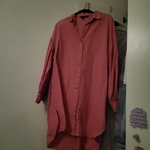 Rosa skjortklänning - Säljer en rosa skjortklänning med knappar framtill. Klänningen har en lös passform och långa ärmar, perfekt för en avslappnad stil. Den är tillverkad i ett lätt och bekvämt material.