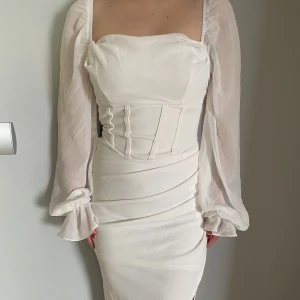 Vit klänning med långa ärmar - Elegant vit klänning med långa, transparenta ärmar och en smickrande slits. Den har en åtsittande passform med en korsettliknande detalj i midjan som ger en fin siluett. Perfekt för speciella tillfällen.