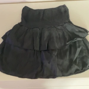 Svart volangkjol - Supersöt svart kjol med volanger och resår i midjan. Kjolen är kort och har en luftig känsla, perfekt för varma dagar. Enkel att matcha med olika toppar för en trendig look. Köpte den i spanien så vet inte vart den är ifrån!