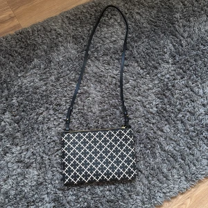 Svart axelväska med vitt rutmönster från By Marlene Birger - Snygg svart axelväska från By Marlene Birger med vitt rutmönster och guldfärgad dragkedja. Väskan har justerbart axelband och är perfekt för att bära det viktigaste. Klassisk och stilren design som passar till många tillfällen. Köpt på Nk för 900 aldrig använd prislapp borttagen för jag trodde jag skulle använda den kvitto och påse finns. Bandet kan tas bort.