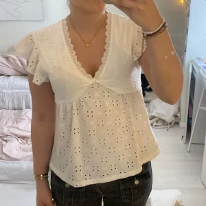 Vit blus med spetsdetaljer - Charmig vit blus med v-ringning och spetsdetaljer. Blusen har korta ärmar med volang och ett broderat blommönster. Perfekt för en somrig look.