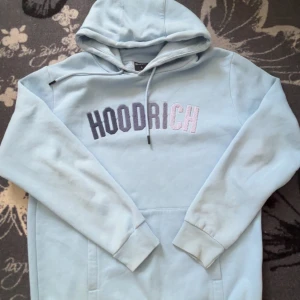 Ljusblå hoodie från Hoodrich - Säljer en snygg ljusblå hoodie från Hoodrich med broderad text på bröstet. Tröjan har en klassisk känguruficka och justerbar huva med dragsko. Perfekt för en avslappnad stil.
