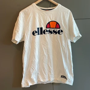 Vit t-shirt från Ellesse - Säljer en klassisk vit t-shirt från Ellesse med deras ikoniska logga i orange och marinblått på bröstet. T-shirten är kortärmad och tillverkad i bomull, perfekt för en avslappnad stil. Passar bra till jeans eller shorts för en casual look.
