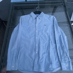 Ljusblå skjorta från Ralph Lauren - Klassisk ljusblå skjorta från Ralph Lauren med button-down krage och broderad logga på bröstet. Skjortan har långa ärmar och knäppning framtill. Perfekt för en stilren och tidlös look. (ÖPPEN FÖR BUD)