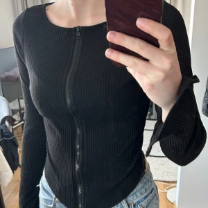Svart ribbad kofta med dragkedja - Säljer en svart ribbad kofta med dragkedja framtill. Tröjan har lång ärm och är figurnära i modellen. Perfekt till jeans eller kjol för en stilren look.