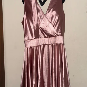 Rosa axelbandslös klänning - Elegant rosa klänning i satin med omlott-design och v-ringning. Axelbandslös stil med ett brett midjeband som ger en smickrande passform. Perfekt för festliga tillfällen.