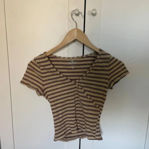 Randig ribbad topp från Hollister - Säljer en beige och vinröd randig ribbad topp från Hollister. Toppen har en v-ringad design och korta ärmar, perfekt för en avslappnad stil. Den är i en slim passform och har en omlott-look framtill.