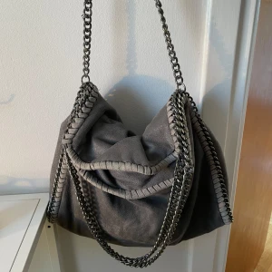 Grå axelväska med kedjedetaljer - Snygg grå axelväska med kedjedetaljer som ger en cool look. Väskan har en mjuk form och är perfekt för att bära med sig det nödvändigaste. Den har en unik design med kedjor som löper längs kanterna och axelremmen.