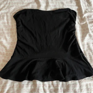 Svart axelbandslös topp - Säljer en svart axelbandslös topp med utsvängd volanger nertill. Enkel och stilren design. Toppen har en figurnära passform upptill och är tillverkad i ett mjukt material.