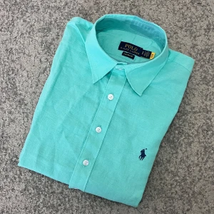turkos skjorta från Polo Ralph Lauren  - Snygg turkos skjorta från Polo Ralph Lauren med klassisk krage och knappar framtill. Skjortan har en broderad logotyp på bröstet och är perfekt för en stilren look.