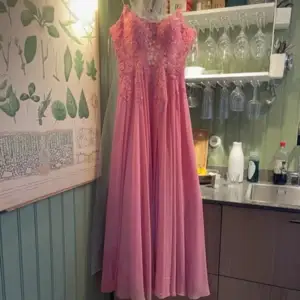 Elegant rosa långklänning med spetsdetaljer och smala axelband. Klänningen har en slits framtill och snörning i ryggen för en perfekt passform. Perfekt för speciella tillfällen.