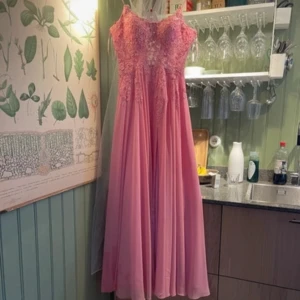 Rosa  balklänning  med spetsdetaljer - Elegant rosa långklänning med spetsdetaljer och smala axelband. Klänningen har en slits framtill och snörning i ryggen för en perfekt passform. Perfekt för speciella tillfällen.