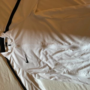 Vit träningströja från Nike - Säljer en vit träningströja från Nike med svarta detaljer och dragkedja. Jackan har långa ärmar och är tillverkad i Dri-FIT-material för optimal komfort under träning. Perfekt för sportiga aktiviteter! Skön och snygg, otroligt skick 