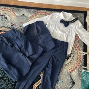 Mörkblå kostym med väst, vit skjorta,byxor och fluga från H&M - Säljer en stilren mörkblå kostym med matchande väst, vit skjorta och en elegant mönstrad fluga. Kostymen har klassisk passform och kavajen har knappar framtill. Perfekt för bal eller fest. För barn. 