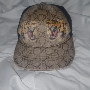 Beige keps med tigertryck från Gucci - Säljer en beige keps från Gucci med klassiskt GG-mönster och två tigrar framtill. Kepsen har svart mesh på sidorna och baktill samt justerbart band med gröna och röda ränder. Perfekt för dig som gillar statement-plagg.