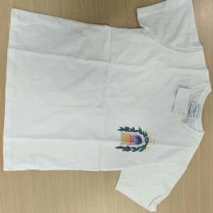 Vit t-shirt med tryck från Casablanca - Vit t-shirt från Casablanca med färgglatt tryck av byggnad och blad på både bröstet och ryggen. T-shirten har rund hals och korta ärmar. Perfekt för dig som gillar unika prints och stilrena plagg.