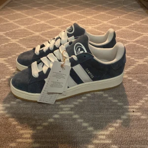 Adidas Campus sneakers i blå mocka - Säljer ett par helt nya Adidas Campus sneakers i blå mocka med vita detaljer och klassiska tre ränder på sidan. Skorna har vit sula och vita skosnören, samt Adidas-logga på plösen och insidan. Perfekta för en stilren vardagslook.