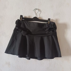 Svart kortkjol med rosetter - Snygg svart kortkjol med söta rosetter i midjan. Kjolen har en elastisk midja för extra komfort och en lätt utsvängd form som ger en fin siluett. Perfekt för att ge en stilren touch till din outfit.