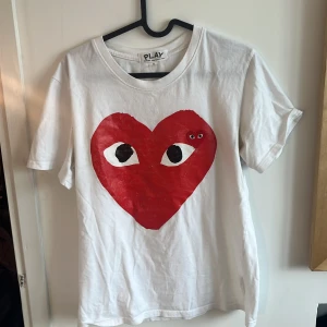 Vit t-shirt från Comme des Garçons - Säljer en vit t-shirt från Comme des Garçons med det ikoniska röda hjärtat med ögon på framsidan. T-shirten är kortärmad och har en normal passform. Perfekt för dig som gillar stilren design med en twist. 🖤.                            Dock sliten därav priset.
