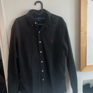 Svart skjorta från Ralph Lauren - Snygg svart skjorta från Ralph Lauren med klassisk krage och knappar framtill. Den har en broderad logotyp i rött på bröstet och är i slim fit. Perfekt för en stilren look.