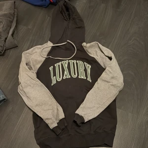 Svart och grå hoodie från Vailent med tryck - Säljer en svart hoodie från Vailent med grå ärmar och stort tryck 'LUXURY' på bröstet. Tröjan har huva med dragsko och muddar vid ärmslut och nederkant. Använd max 3 gånger då den aldrig riktigt varit min stil. Men den är väldigt fin och mjuk 