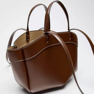 Brun handväska - Slutsåld väska från Zara, aldrig använd. Stilren brun handväska med kontrastsömmar och två handtag. Väskan har ett rymligt huvudfack och ett löstagbart innerfack med dragkedja.