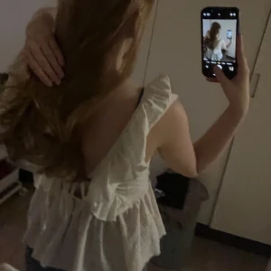 Vit ärmlös blus med volang - Säljer en söt vit blus med volangkant och broderade detaljer. Toppen är ärmlös med breda axelband och har en lätt utsvängd peplumform. Perfekt för varma dagar och enkel att matcha med jeans eller kjol.