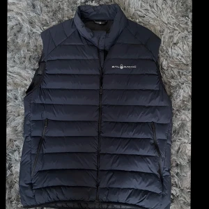 Svart dunväst från Sail Racing - Snygg svart dunväst från Sail Racing med dragkedja och logotyp på bröstet. Perfekt för kyliga dagar med sin quiltade design och praktiska fickor.