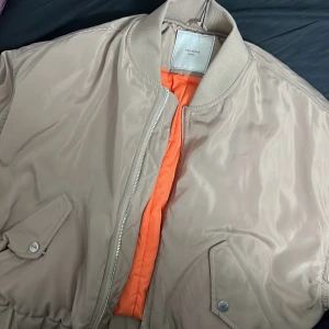 Beige bomberjacka från Neo Noir - Snygg beige bomberjacka från Neo Noir med orange foder. Jackan har dragkedja framtill och två fickor med knappar. Perfekt för en cool och avslappnad stil. Den är helt ny men jag har bytt stil priset går att diskutera vid snabb affär
