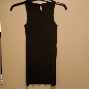 Svart ribbad kortklänning POMPdeLUX - Svart ärmlös kortklänning från POMPdeLUX i ribbad bomull. Klänningen har fina spetsdetaljer längs kanterna och en rak, slim passform. Perfekt för dig som gillar enkel men snygg stil. Det finns ett litet hål längst ner på klänningen vid sidan av sömmen, de e bara att skriva DM för fler bilder