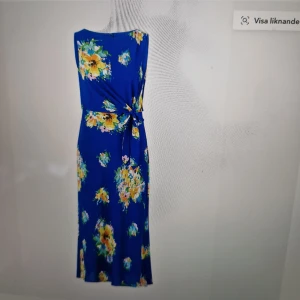 Blommig blå långklänning med knyt - Superfin långklänning i starkt blått med färgglada blommor i gult, grönt och rosa. Ärmlös modell med knyt i midjan som ger en snygg siluett. Perfekt för sommarens alla tillfällen när du vill sticka ut lite extra.