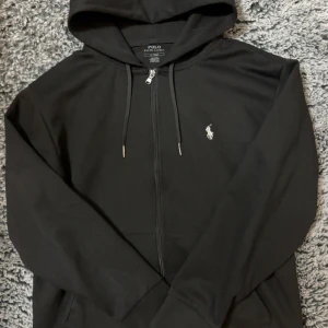 Svart hoodie från Polo Ralph Lauren - Säljer en stilren svart hoodie från Polo Ralph Lauren. Den har en dragkedja framtill och en liten broderad logga på bröstet. Perfekt för både höst och vinter, med en skön huva och långa ärmar. Passar till alla tillfällen!