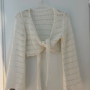 Vit stickad omlottkofta med knytband - Säljer en superfin vit stickad omlottkofta med långa ärmar och knytband framtill. Koftan är croppad och har ett luftigt, öppet stickat mönster som ger en chill och trendig vibe. Perfekt att slänga över ett linne för en snygg lager-på-lager-look.