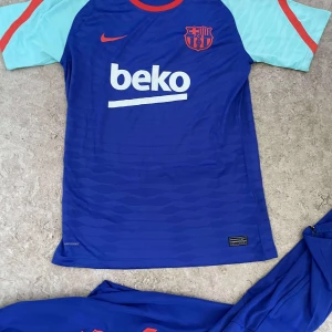 FC Barcelona Nike blå fotbollströja - FC Barcelona träningströja från Nike i blått med ljusblå ärmar och röda detaljer. Tröjan har klubbmärke och sponsortryck på bröstet samt rund halsringning. Tillverkad i lätt och ventilerande funktionsmaterial, perfekt för fotbollsträning.