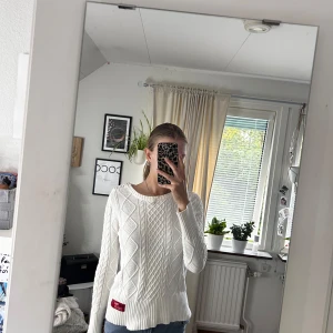 Vit stickad tröja från MarinAlpin - Vit stickad tröja från MarinAlpin med grovt kabelstickat mönster och ribbade muddar. Rund halsringning och dekorativa knappar på ena axeln. Snygg detalj med röd logolapp nertill. Perfekt till jeans för en clean look.