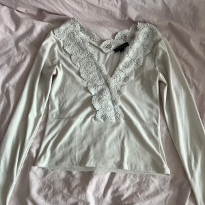 Vit långärmad blus med spets - Supersöt vit långärmad blus med djup v-ringning och breda spetsdetaljer framtill och längs ryggen. Toppens material är mjukt och lätt, perfekt för dig som gillar romantiska vibes och snygga detaljer.